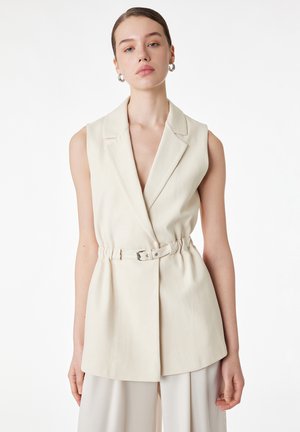 Frau trägt ärmellosen beigen Blazer mit Gürtel, steht vor weißem Hintergrund und schaut mit neutralem Ausdruck nach vorne.