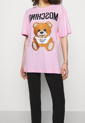 Osoba nosząca różową, oversize'ową koszulę z brązowym misiem i odwróconym napisem "MOSCHINO", zestawioną z czarnymi spodniami.