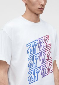 Hombre con camiseta blanca de manga corta con patrón geométrico azul y rojo parecido a un laberinto en el pecho contra un fondo liso.