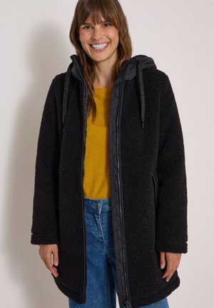 Femme souriante portant une veste polaire noire zippée avec capuche, un t-shirt jaune moutarde et un jean bleu sur un fond neutre.