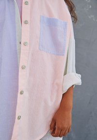 Striped shirt featuring colors orange, lavender, and white, with a pocket on the lavender section and button front closure. Textured fabric.