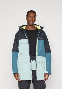 Wodoodporna kurtka w kolorze zielono-niebieskim, z wieloma przednimi kieszeniami, kieszenią na zamek błyskawiczny i jas yellow wnętrzem.