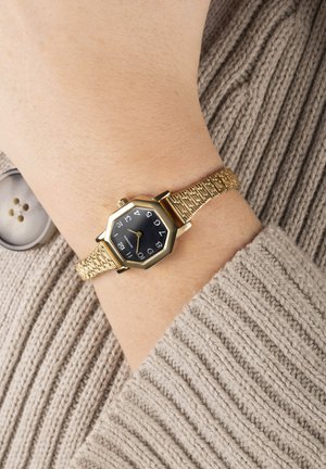 Montre hexagonale dorée avec un cadran noir arborant des chiffres blancs, montée sur un bracelet en maille dorée texturée, affichée sur un poignet.