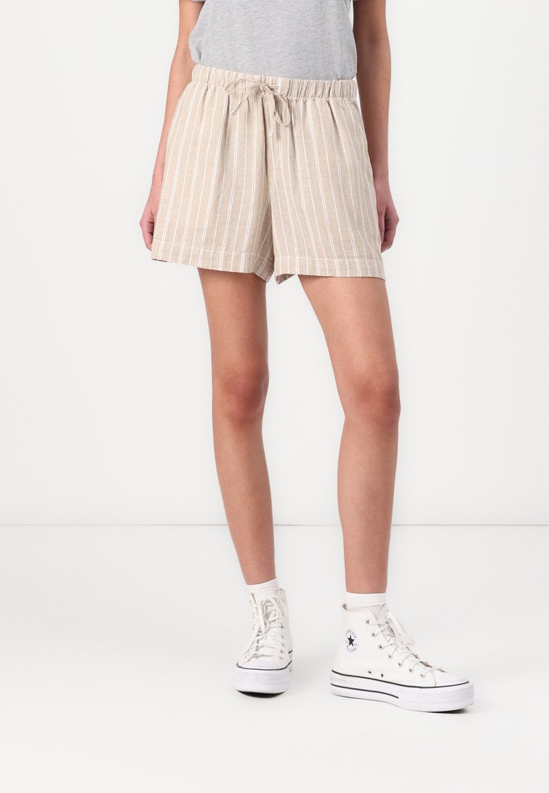 Personne portant un short rayé beige et blanc avec un cordon, un haut gris, des baskets montantes blanches et des chaussettes blanches, debout devant un fond blanc.