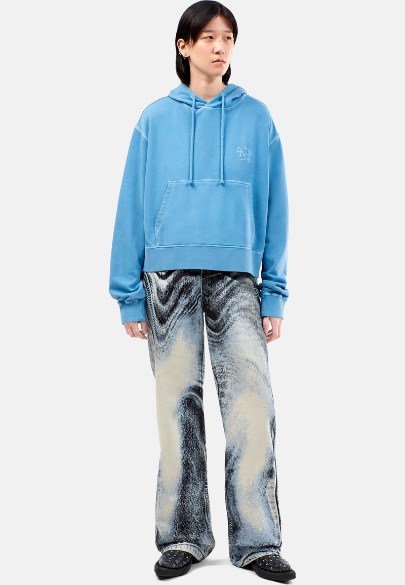 Felpa oversize blu chiaro con tasca frontale; abbinata a jeans a gamba larga con un motivo marmorizzato nero e crema. Calzature nere.
