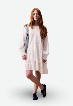 Jeune femme aux longs cheveux roux portant une robe blanche à hauteur des genoux, des baskets noires et un sac à bandoulière bleu clair.
