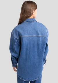 Hele sinine denimmärk, millel on vabaja lõige, terav krae, pikad varrukad ja kumer äär. Nähtavad õmblused.