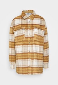 Camisa de franela en un patrón de cuadros amarillos, marrones y blancos. Cuenta con un collar, cierre de botones en la parte delantera y dos bolsillos en el pecho con cierres a presión.