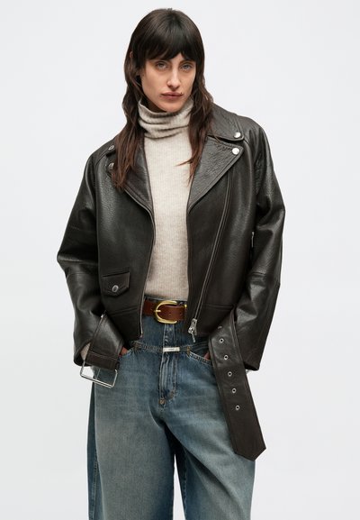 Veste motard en cuir noir avec accessoires argentés, fermeture éclair et col cranté. Portée sur un col roulé beige et un jean taille haute.