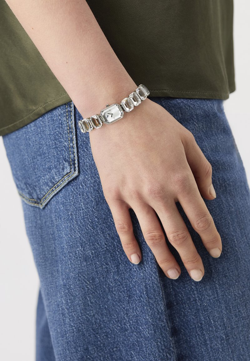 Silver bracelet watch featuring an octagonal face, clear crystal links, and a simple white dial with black markings. Positioned on a wrist.