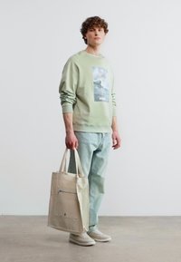 Νέος άνδρας που φοράει ανοιχτό πράσινο φούτερ και τζιν, κρατώντας μια μπεζ τσάντα tote, στέκεται σε τσιμεντένιο πάτωμα μπροστά από έναν λευκό τοίχο.