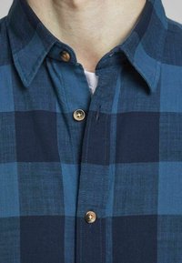 Närbild av en person som bär en blå och mörkblå rutmönstrad skjorta över en vit t-shirt.