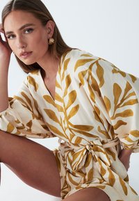 Lange mouw wikkeljurk met een crèmekleurige stof en mosterdgele bladpatronen, getailleerd met een bijpassende riem. Gestructureerd, lichtgewicht materiaal.