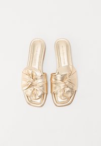 Sandales dorées en simili cuir avec un design à l'avant noué, bout carré et semelle plate. Texture lisse avec un léger éclat.