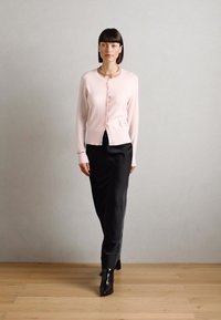 Lys rosa cardigan med en tettsittende design og knappfeste, kombinert med et svart, flytende, ankel-langt skjørt og svarte ankelstøvler.