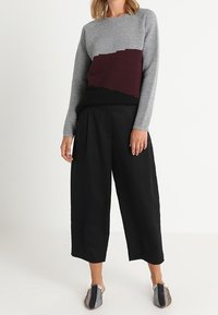 Pull gris côtelé avec accent bordeaux, associé à un pantalon noir à jambes larges et des chaussures à enfiler à motif. Texture lisse sur toute la surface.