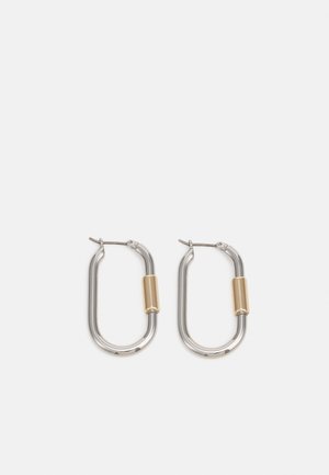 Boucles d'oreilles ovales en argent avec un petit accent cylindrique en or sur chacune, présentées sur un fond clair uni.