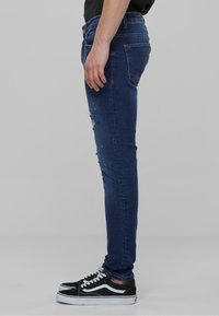 Jeans ajustados de color azul con detalles rasgados en el muslo, combinados con zapatillas negras con acentos blancos. Bucles de cinturón visibles y bolsillos.