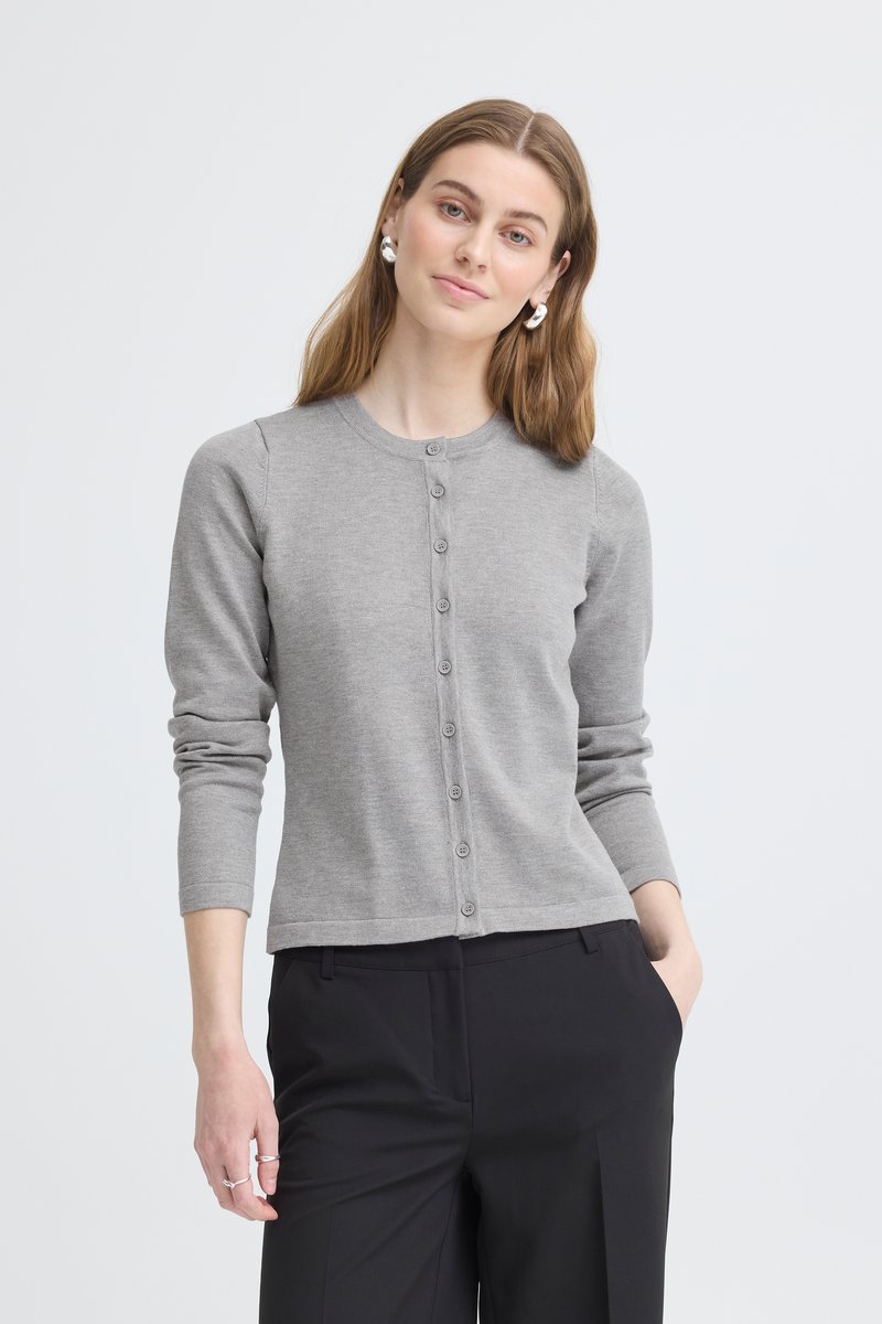 Femme aux cheveux châtain clair portant un cardigan gris boutonné et un pantalon noir, une main dans la poche, debout devant un fond blanc.