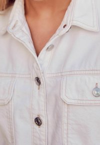 Camicia di jeans bianchi con colletto classico, bottoni argentati e due tasche sul petto con cuciture decorative. Texture morbida e design strutturato.