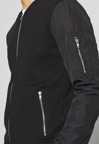 Chaqueta bomber negra con cuerpo de lana y mangas de nailon liso, bolsillos con cremallera y cierres metálicos.