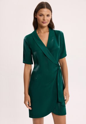 Kobieta o brązowych włosach, ubrana w ciemnozieloną, satynową sukienkę kopertową z krótkim rękawem, głębokim dekoltem w kształcie litery V i wiązaniem po bokach.