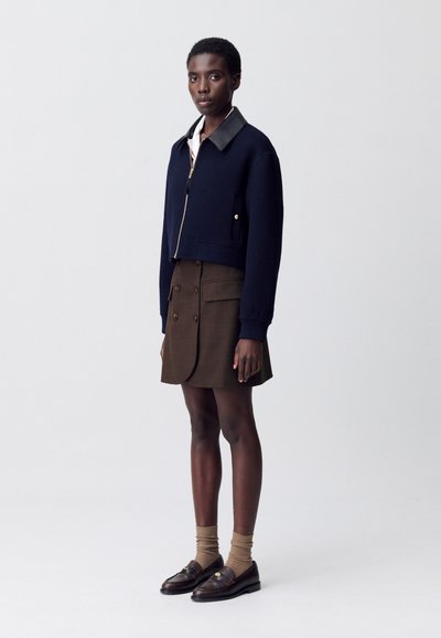 Giacca cropped blu navy con colletto nero, zip e dettagli dorati; gonna marrone con bottoni, abbinata a mocassini scuri e calze marroni.