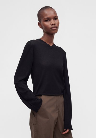 Jeune personne aux cheveux courts portant un haut noir à manches longues et un pantalon marron, une main dans la poche, sur un fond uni.