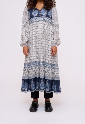 Femme portant une robe blanche à manches longues avec des motifs floraux bleus, un pantalon noir court et des mocassins noirs sur un fond neutre.