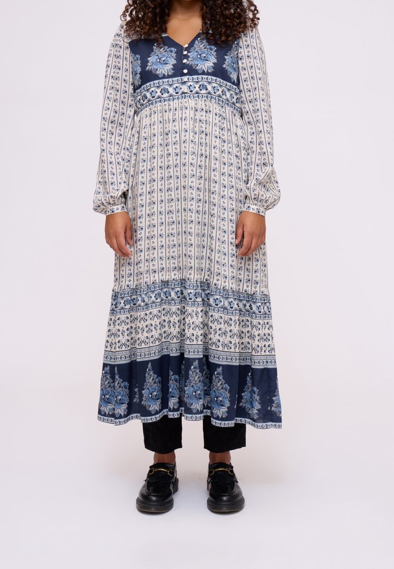 Femme portant une robe blanche à manches longues avec des motifs floraux bleus, un pantalon noir court et des mocassins noirs sur un fond neutre.