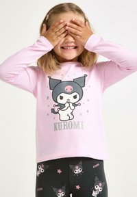 Lachend kind bedekt zijn ogen met handen, draagt een roze shirt en donkere legging met Kuromi karakters en tekst.