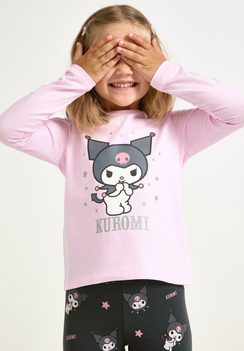 Lachend kind bedekt zijn ogen met handen, draagt een roze shirt en donkere legging met Kuromi karakters en tekst.