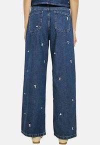 Platas kājas zilie džinsi no denima ar izšūtām ziedu detaļām, piecu kabatu dizains un neapstrādāta apdare, demonstrējot brīvu griezumu un ikdienas tekstūru.