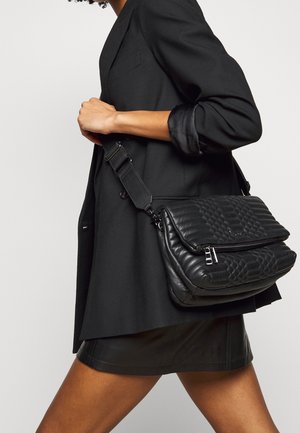 Persona llevando un bolso bandolera de cuero acolchado negro con cremallera, bajo una chaqueta negra con la manga enrollada y una falda corta negra.