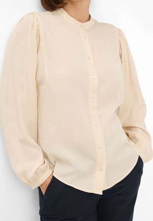 Femme portant un chemisier beige boutonné avec de longues manches bouffantes et un pantalon noir, mains dans les poches, sur un fond blanc uni.