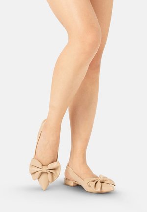 Gołe nogi w beżowych płaskich butach z dużymi ozdobnymi kokardami, jedna stopa lekko uniesiona na palcach na jednolitym tle.