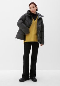 Czarna kurtka puchowa z kapturem, zestawiona z musztardowym swetrem w serek, czarnymi spodniami dzwonami i czarnymi masywnymi butami. Jednolita tło.