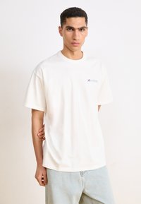 Hvid bomulds T-shirt med afslappet pasform, korte ærmer og rund hals. Har et farverigt logo på venstre bryst. Et par lyseblå bukser.