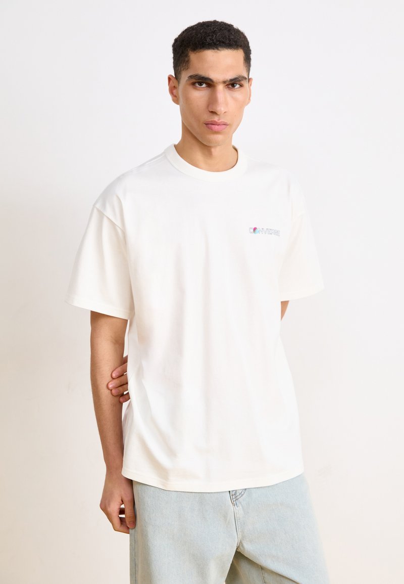 Hvid bomulds T-shirt med afslappet pasform, korte ærmer og rund hals. Har et farverigt logo på venstre bryst. Et par lyseblå bukser.