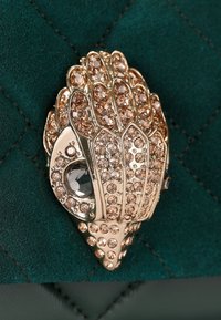 Sfondo in tessuto trapuntato verde con una chiusura decorativa dorata caratterizzata da numerosi strass color rosa e una gemma centrale trasparente.