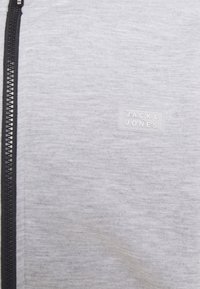 Tissu gris clair avec une fermeture éclair noire sur la gauche et un petit écusson rectangulaire du logo "Jack & Jones" sur le côté droit.