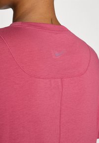 Vaaleanpunainen Nike-t-paita, jossa on röyhelömäinen pyöreä kaula-aukko ja hienovaraiset ompeluyksityiskohdat. Selässä pienempi vaalea violetti logo.