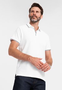 Vit polo-shirt med krage, med marinblå kant vid kragen och ärmarna. Tyget har en strukturerad yta.