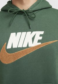 Felpa verde con tasca a marsupio, caratterizzata da un logo "NIKE" bianco e uno swoosh marrone testurizzato. Cappuccio con cordino e polsini a coste.