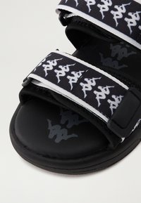Sorte slidesandal med justerbare stropper. Stropper har et hvitt logomønster. Sålen er laget av teksturert svart materiale som gir godt grep.
