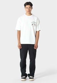 Hvid t-shirt med korte ærmer og grafisk print på brystet, sammen med sorte vide bukser og sorte sneakers med hvide såler.