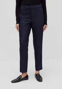 Marineblauwe pantalon met een gladde, iets glanzende textuur, met een standaard taille, rechte benen en zijzakken. Draagt met zwarte instapschoenen.