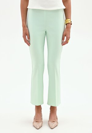 Femme portant un pantalon ajusté vert menthe, un haut blanc, des talons pointus beiges et des bijoux en or au poignet gauche et à la main droite.
