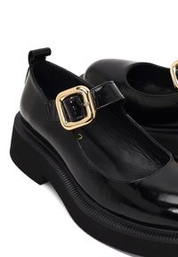 Schwarze Lackleder-Mary-Jane-Schuhe mit runder Zehenform, goldener Schnalle und chunky schwarzer Sohle mit geriffelter Detailierung.