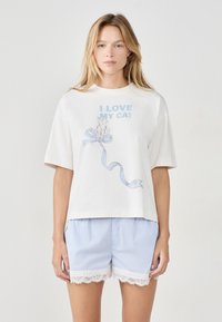 Mujer rubia vistiendo una camiseta blanca con el texto "AMO A MI GATO" y una ilustración de un gato, combinada con pantalones cortos a rayas azul claro con encaje.